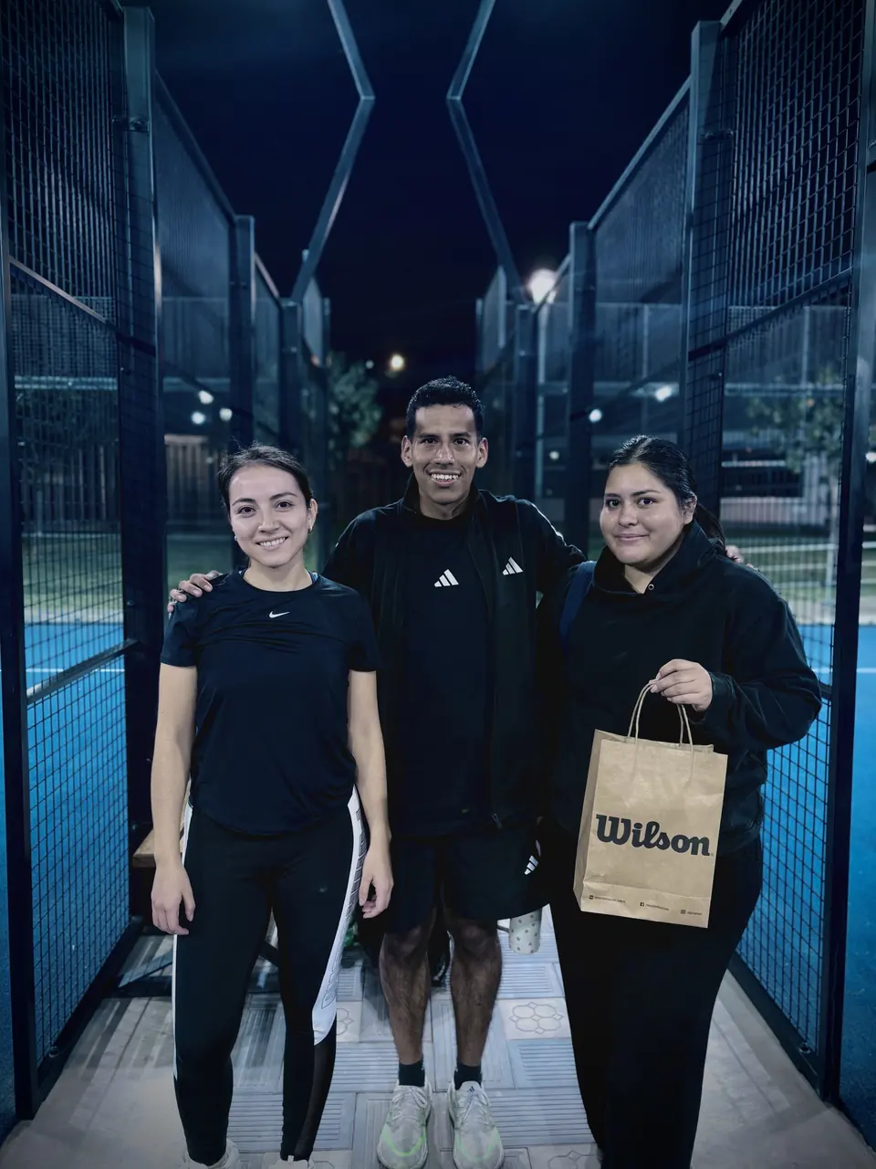 Tres jugadores sonriendo en el pasillo de una cancha de pádel de noche