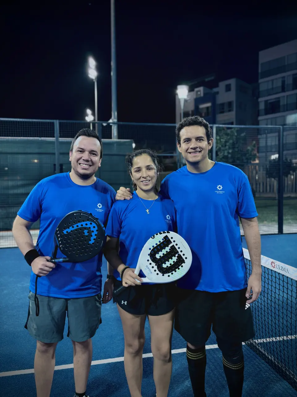 Tres jugadores con polos azules posando junto a la red de una cancha de noche