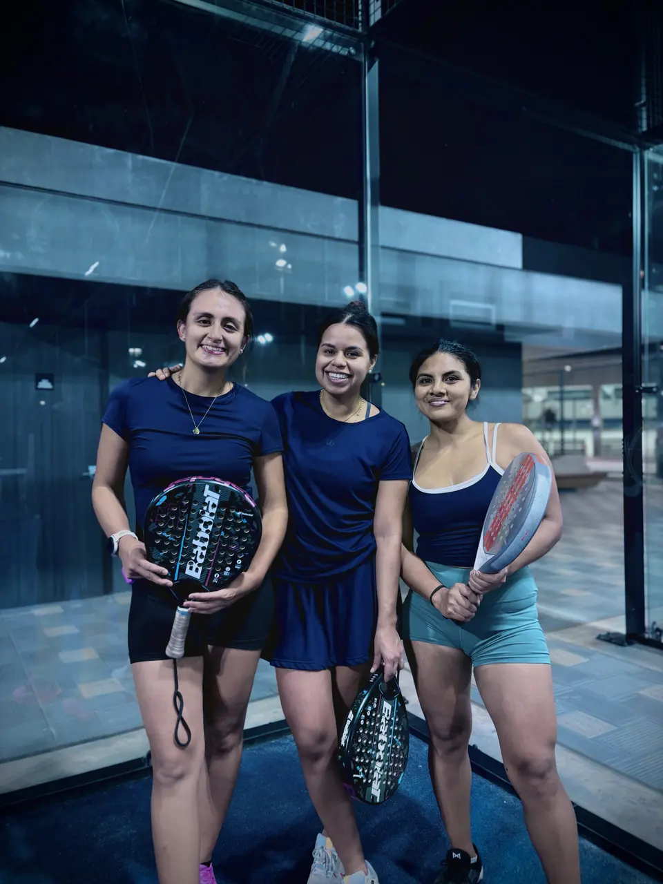 Tres jugadoras sonriendo con sus raquetas después de jugar de noche